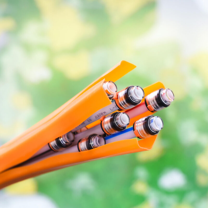 Nahaufnahme von orangefarbenen Röhren, die mehrere Glasfaserkabel umhüllen. Die Kabel enden in silberfarbenen Steckverbindungen. Im Hintergrund sind verschwommene, grüne und gelbe Farben.