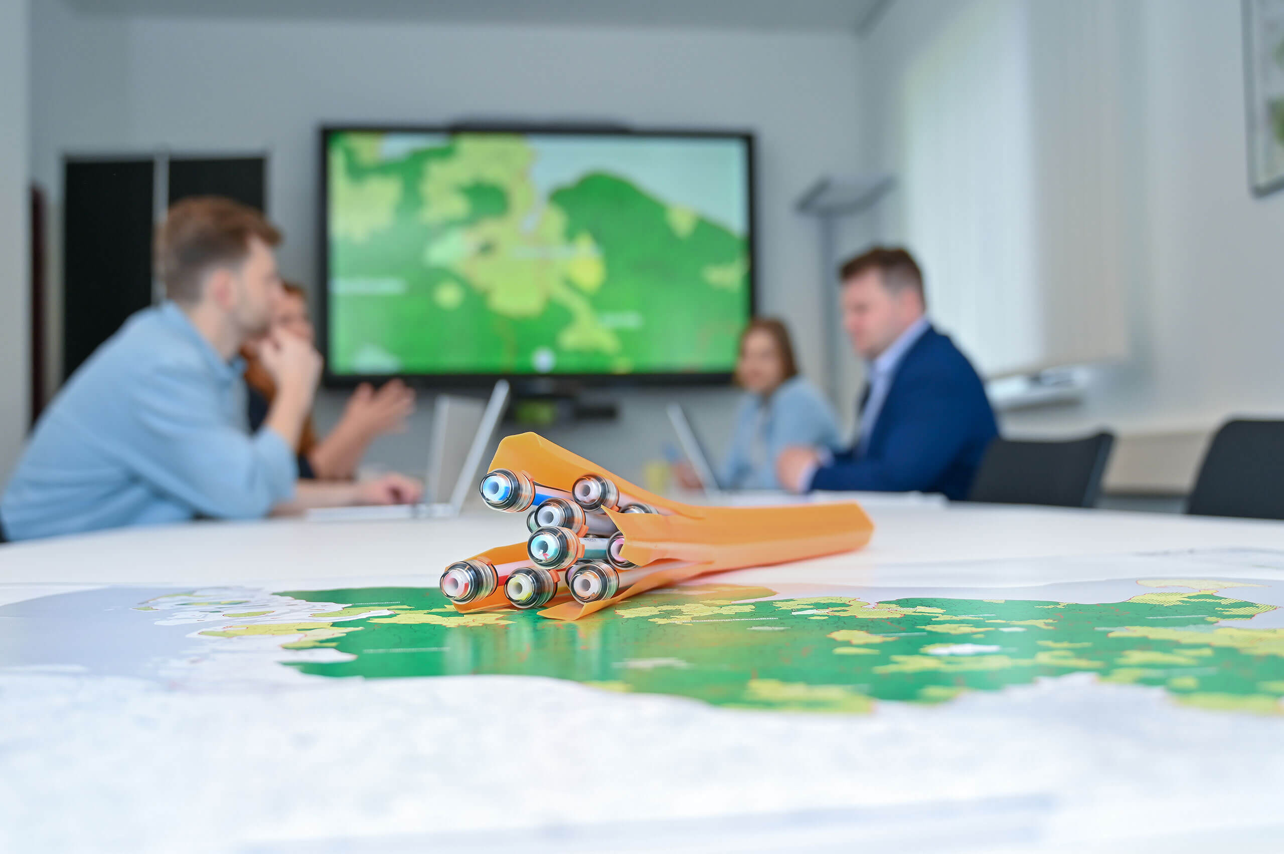 Meernetz Sh Verfuegbarkeitscheck 3 Ein Besprechungsraum mit vier Personen im Hintergrund, die an einem Tisch sitzen und diskutieren. Im Vordergrund liegt eine gefaltete Landkarte und eine Packung mit Buntstiften. Ein Bildschirm zeigt eine Landkarte.