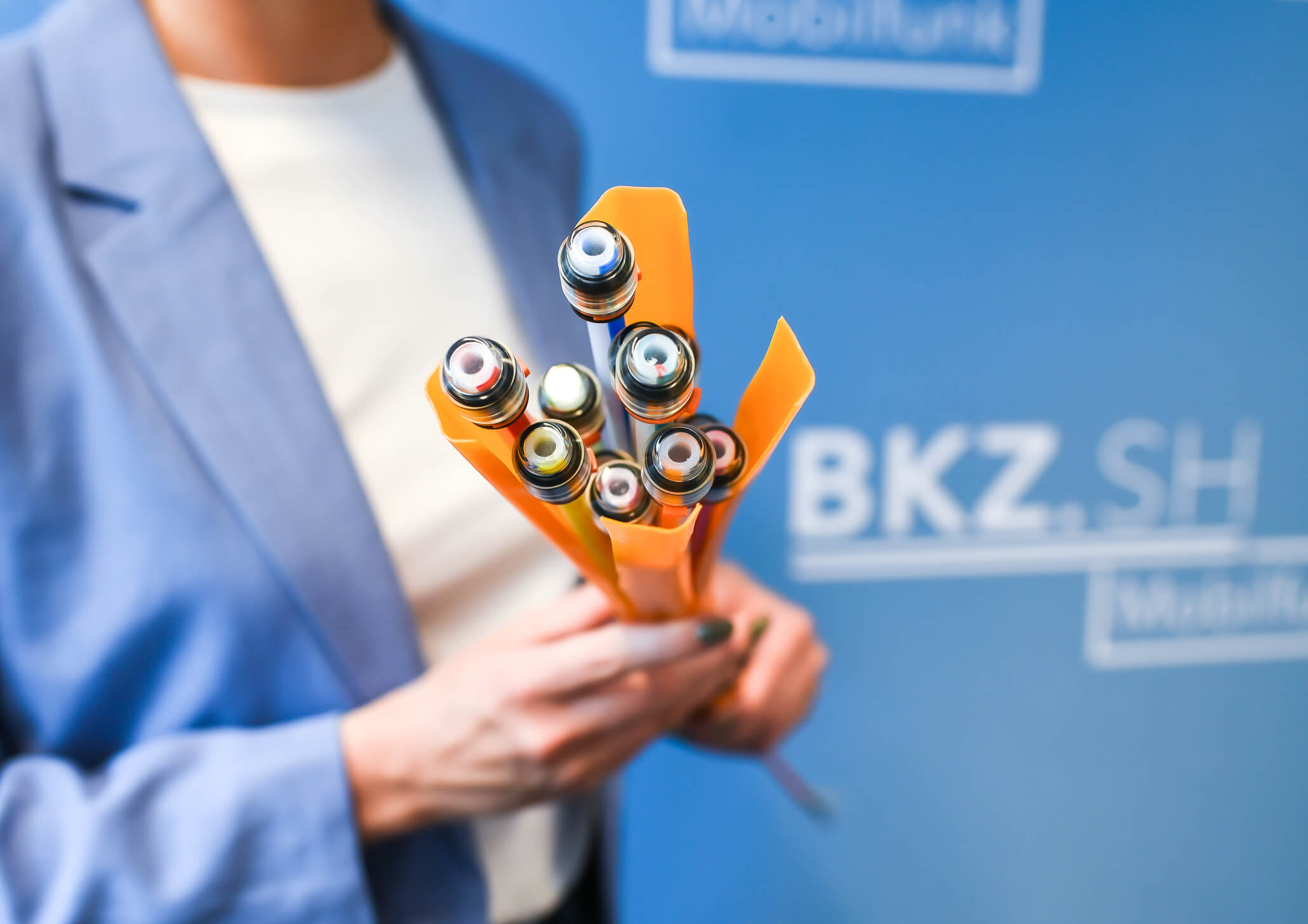 Meernetz Sh Warum Glasfaser Eine Person hält ein Bündel moderner Glasfaserkabel, die aus einer orangefarbenen Ummantelung ragen. Der Hintergrund ist blau mit weißen, unscharfen Schriftzügen. Die Person trägt einen blauen Blazer und ein weißes Oberteil.