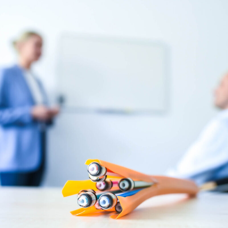 Im Vordergrund sind bunte Netzwerk- oder Glasfaserkabel zu sehen. Im unscharfen Hintergrund stehen eine Person in einem blauen Sakko und eine sitzende Person vor einem leeren Whiteboard.